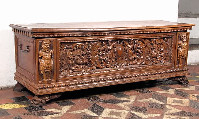 Ornately carved wooden chest featuring classical female figures, scrolling foliage, and detailed relief on tiled floor.